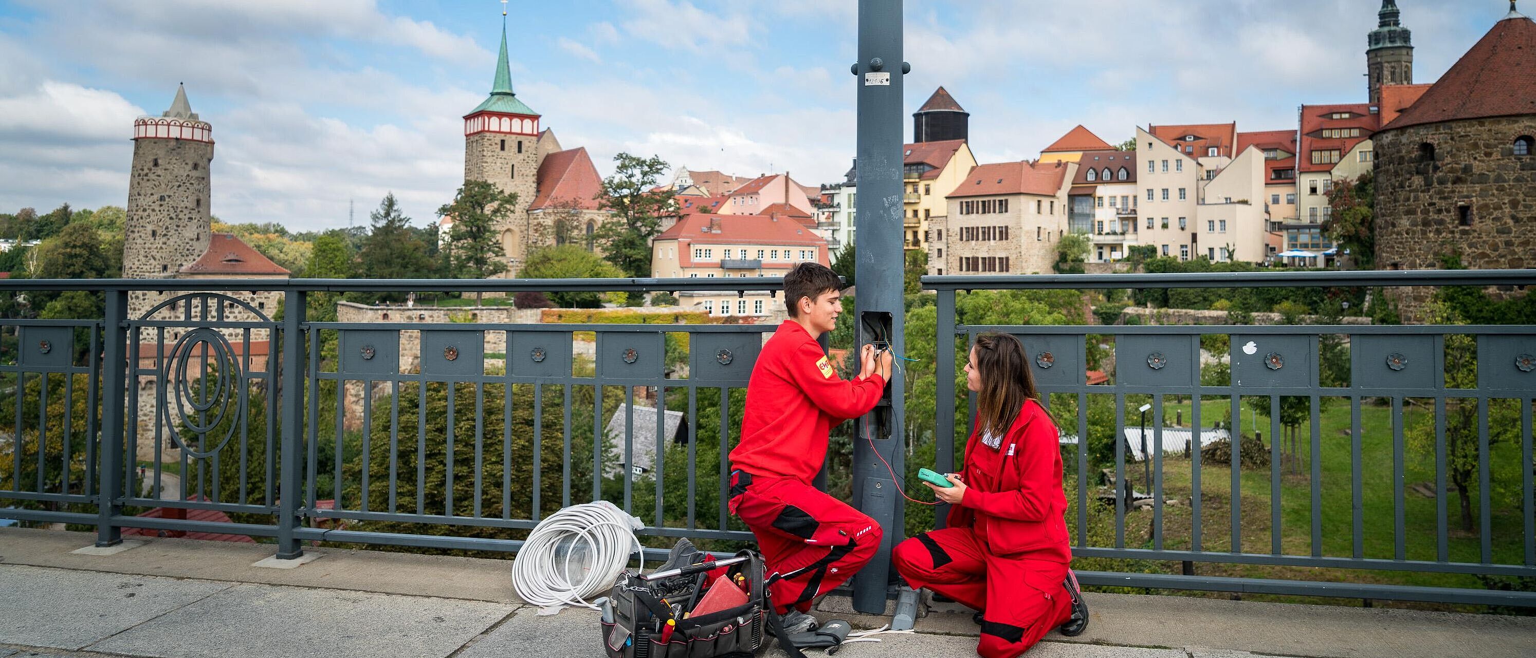 Zwei Personen in Arbeitskleidung eines Elektrotechnikers reparieren eine Laterne auf einer Bautzener Brücke. Im Hintergrund ein Panoramablick auf Bautzen.