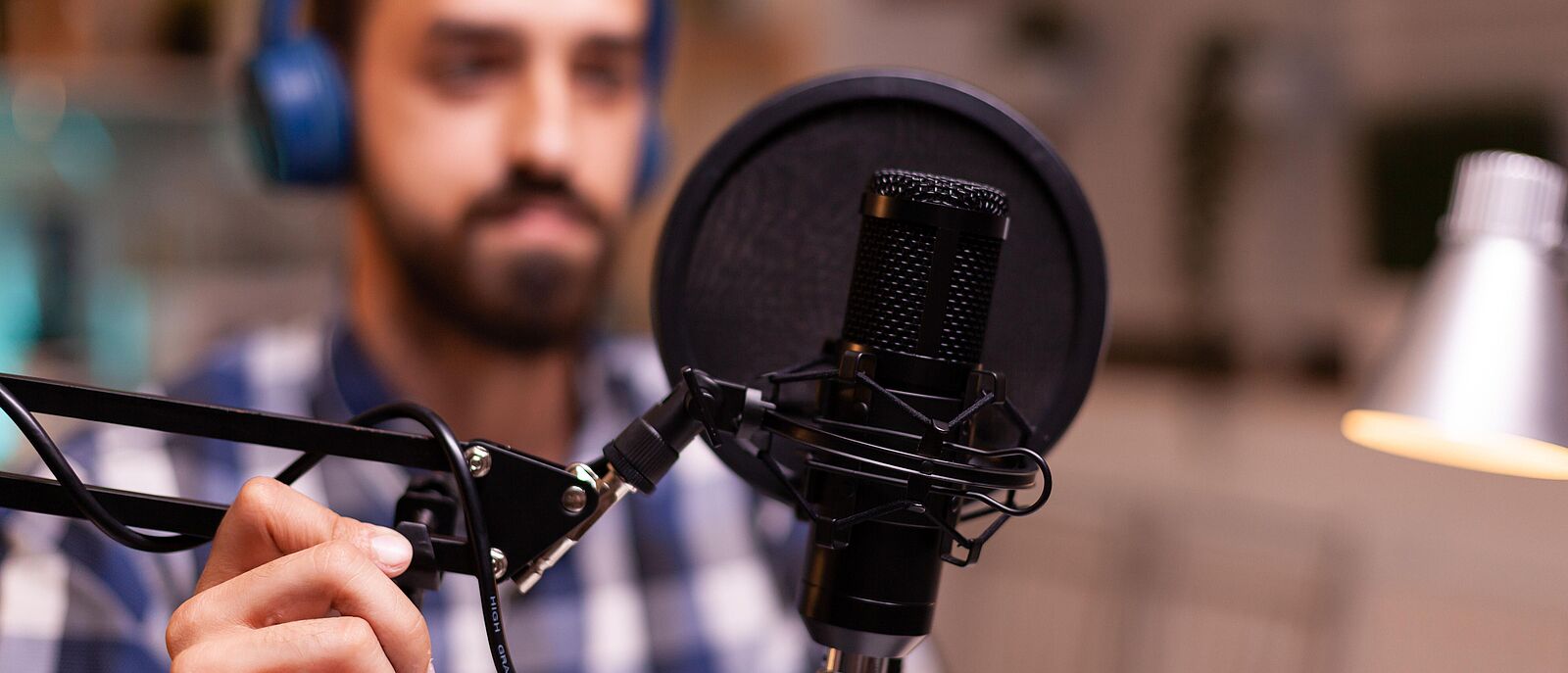 Eine Person sitzt in einem Aufnahmestudio, vor sich ein Aufnahmemikrofon welches er mit der rechten Hand zu sich schiebt.