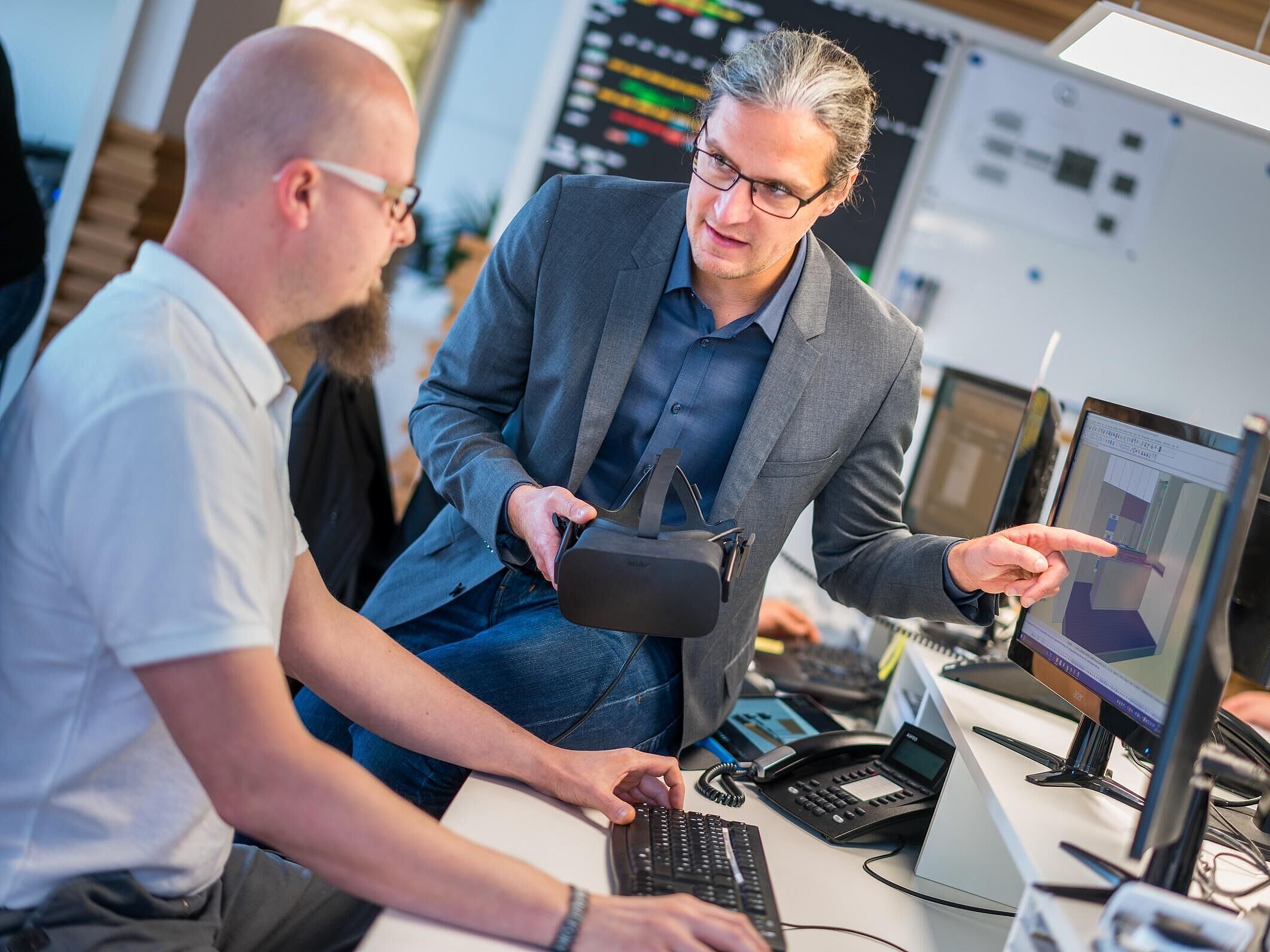 Zwei Personen sitzen an einem Computer und diskutieren ein Projekt. Eine Person hat eine VR-Brille in der Hand.