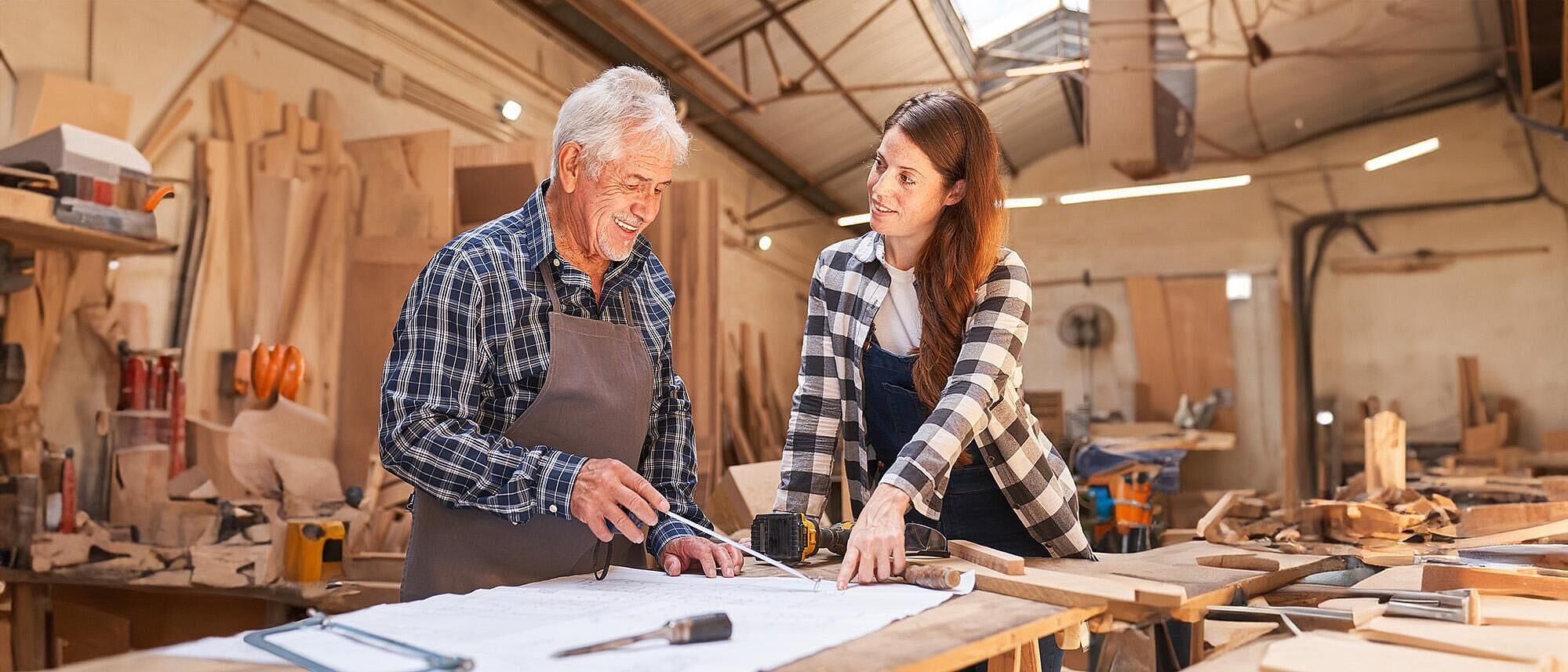 Ein Senior Handwerker steht in einer Tischlerei mit einer weiteren Person in Arbeitskleidung an einem Arbeitstisch, vor sich ein Bauplan den sie sich beide prüfend auswerten. Auf diesem Bauplan liegen verschiedene Werkzeuge, wie Pinsel, Akkuschrauber, Meßband, Holzsäge und zwei Holzlatten in verschiedenen Breiten.