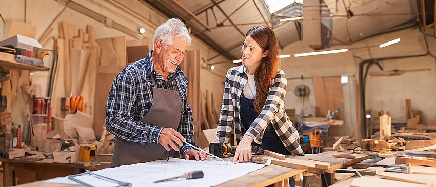 Ein Senior Handwerker steht in einer Tischlerei mit einer weiteren Person in Arbeitskleidung an einem Arbeitstisch, vor sich ein Bauplan den sie sich beide prüfend auswerten. Auf diesem Bauplan liegen verschiedene Werkzeuge, wie Pinsel, Akkuschrauber, Meßband, Holzsäge und zwei Holzlatten in verschiedenen Breiten.