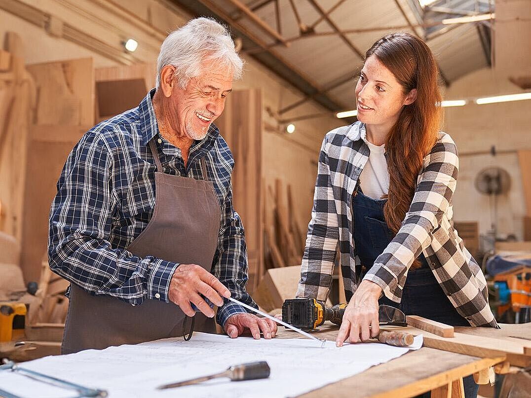 Ein Senior Handwerker steht in einer Tischlerei mit einer weiteren Person in Arbeitskleidung an einem Arbeitstisch, vor sich ein Bauplan den sie sich beide prüfend auswerten. Auf diesem Bauplan liegen verschiedene Werkzeuge, wie Pinsel, Akkuschrauber, Meßband, Holzsäge und zwei Holzlatten in verschiedenen Breiten.