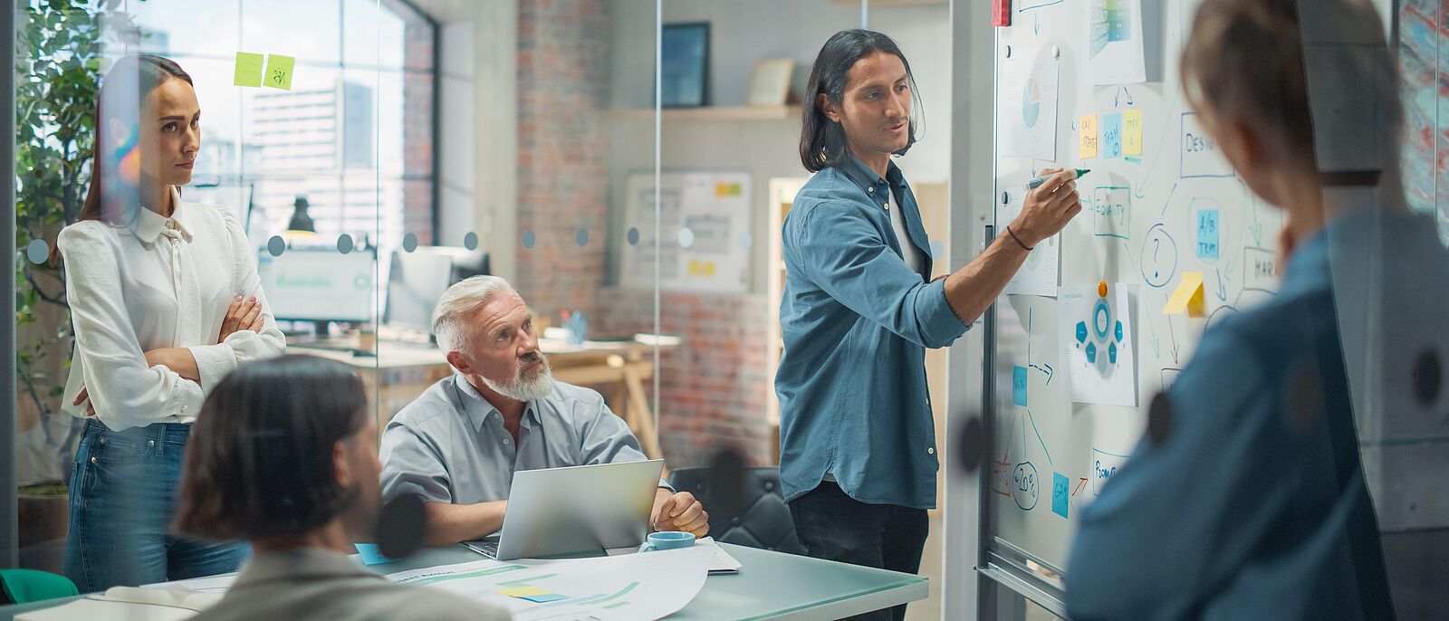 Innovationssprint 2026 – Teilnahmeaufruf In einem Beratungsraum steht ein Vortragender an einer Moderationstafel und schiebt dort angebrachte Moderationskarten hin und her. Mehrere Zuhörer sitzen oder stehen davor an einem Konferenztisch und diskutieren mit dem Referenten.