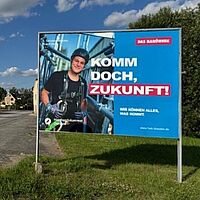 Das Bild zeigt zu zwei Dritteln einen Menschen in Sicherheitsausrüstung, der auf einem hellblauen Hintergrund zu sehen ist und an einen Bergsteiger erinnert. Des Weiteren ist der Spruch \"Komm doch Zukunft\" zu sehen. Darunter der Zusatz \"Wir können alles, was kommt.\"