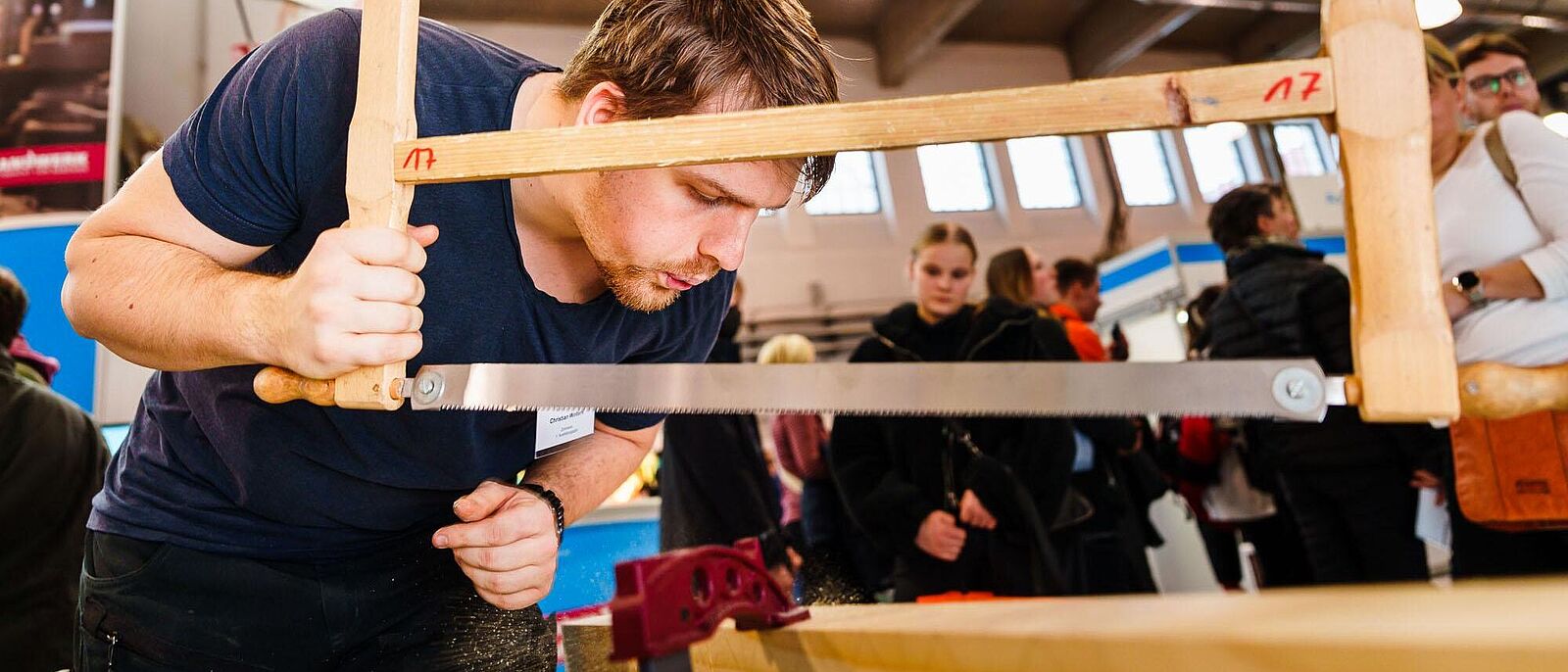 Zimmerer im ersten Ausbildungsjahr, arbeitet an einem Messestand mit einer Holzsäge an einer Seilhieb-Verzierung.