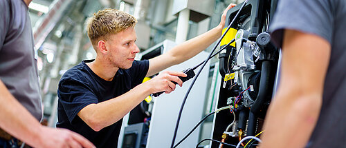 Ein Lehrling führt Arbeiten/Messungen an einem Motor ein einem Montageschrank aus und wird von zwei Ausbildern links und rechts beraten, begleitet