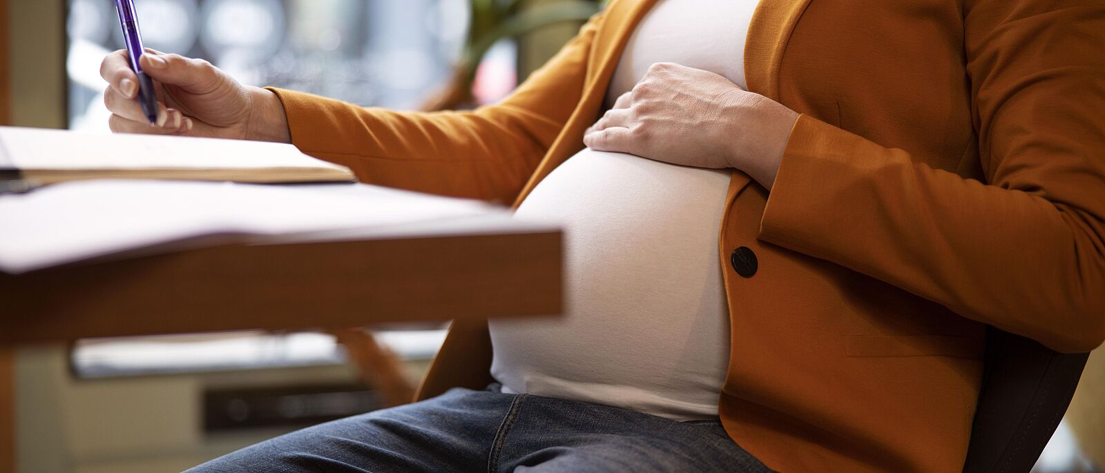 Makroaufnahme: Eine schwangere Frau sitz am Schreibtisch und Dokumente vor sich liegen. Auf einem Dokument notiert sie etwas, dabei liegt ihre linke Hand auf dem Bauch.