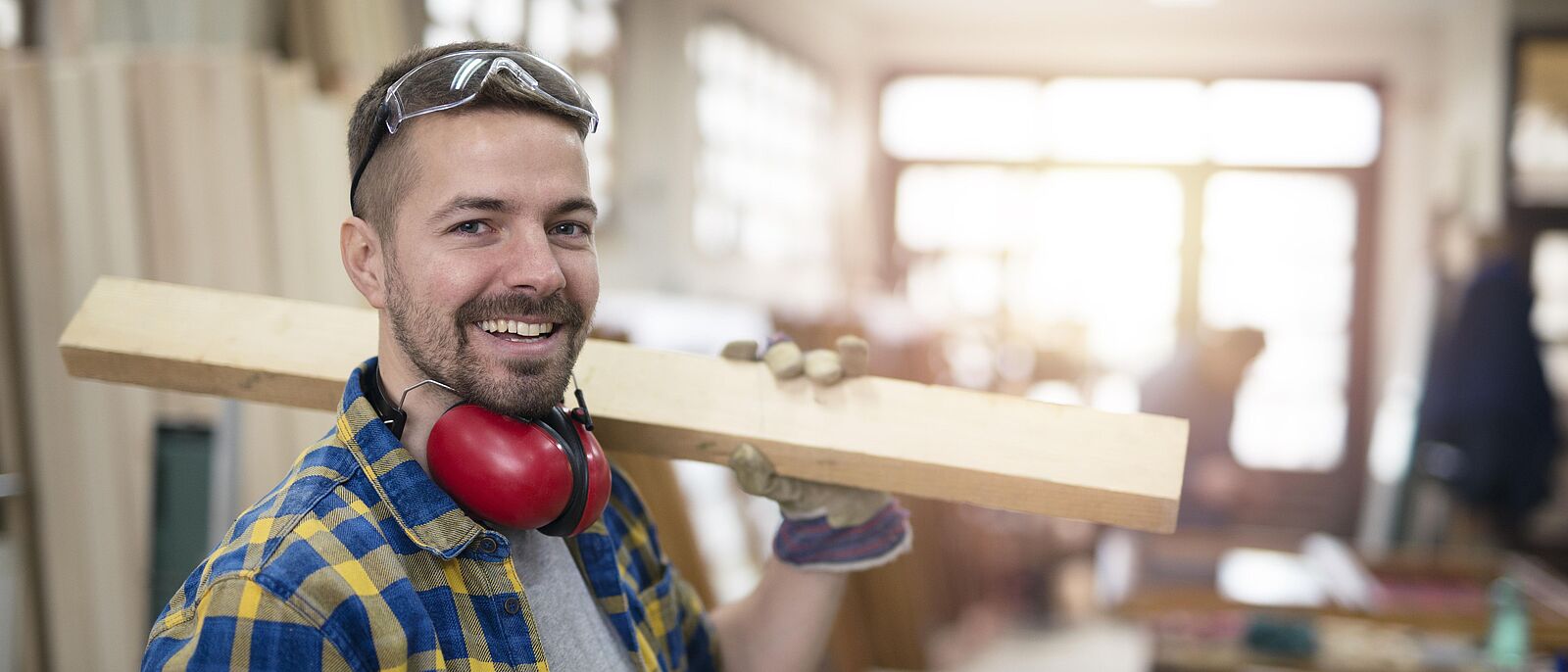 Ein Mann steht mit einem bearbeiteten Holzbrett auf der Schulter und einem Hörlärmschutz um den Hals in einer Werkstatt.