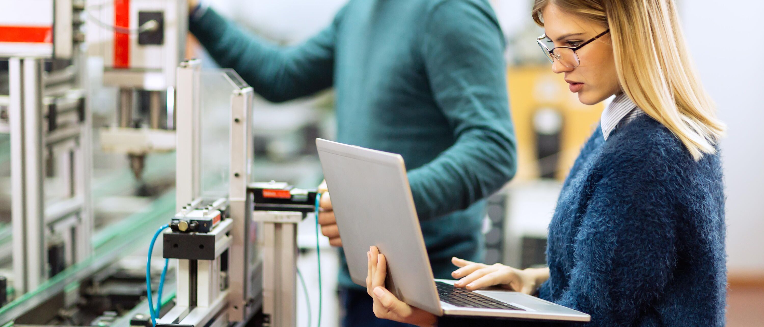 Zwei Personen bauen an einem technischen Modell. Die eine Person schaut in einen Laptop, die andere überprüft an den Einstellungen des technischen Modells etwas.