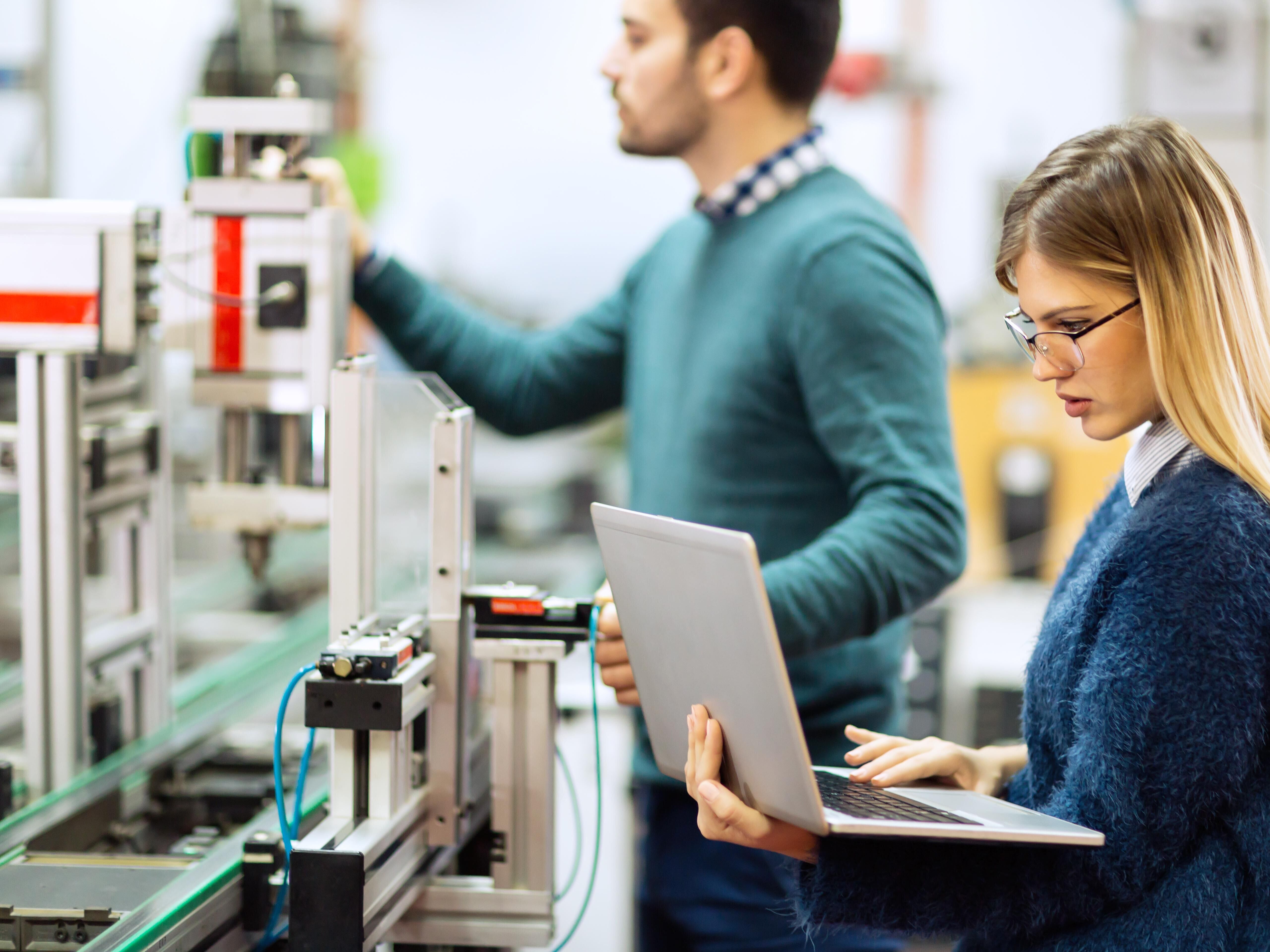 Zwei Personen bauen an einem technischen Modell. Die eine Person schaut in einen Laptop, die andere überprüft an den Einstellungen des technischen Modells etwas.