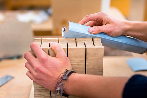 Makroaufnahme: in dermitte auf einer Holzwerkbank steht ein Holzwerkstück, welches eine Person mit einem Schleifklotz bearbeitet.