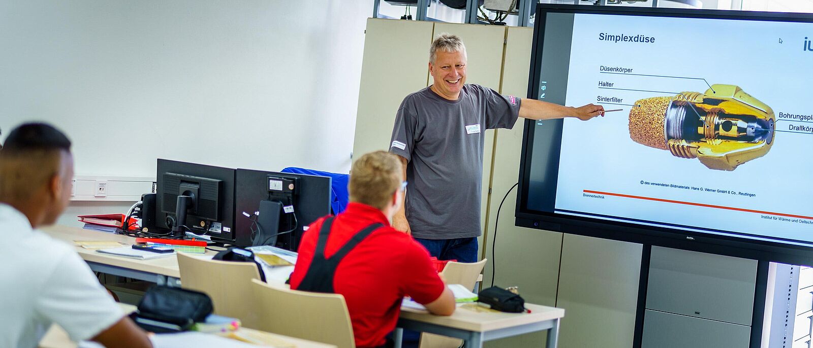 Blick in einem Unterrichtsraum der Metalltechnik. Ein Ausbilder gibt Erläuterungen anhand einer Präsentation an einer interaktiven Tafel. Zwei weitere Personen sitzen mit Unterrichtsmaterial an Unterrichtstischen.