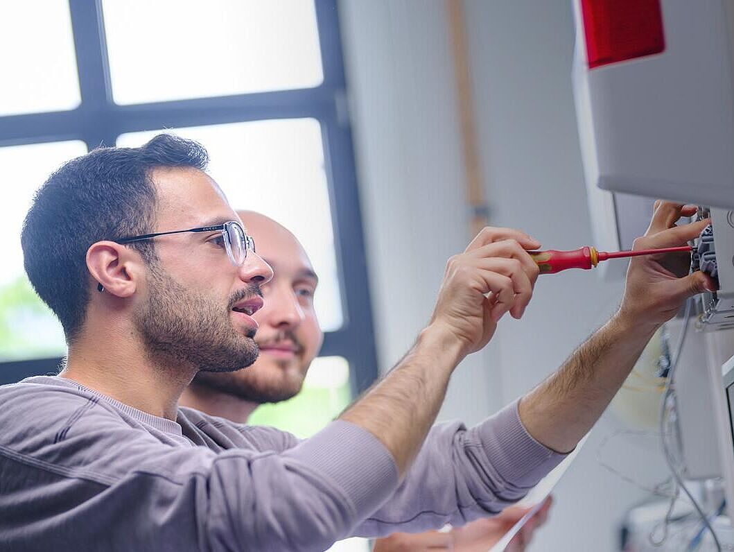 Gesellen- und Abschlussprüfungen Bildcollage einer Person,  die an einem Schulungswerkstattplatz sitzt und an einem Schaltkasten arbeitet. Rechts daneben weißer Text auf blauem Hintergrund \"Wer gute Dinge macht, kann guter Dinge sein.\"