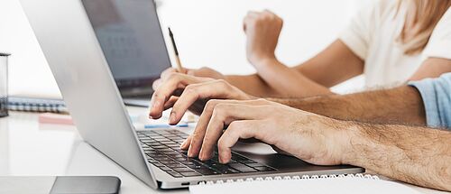 Zwei Personen sitzen zusammen am Schreibtisch, vor sich jeder ein Laptop. Beide sehen in einen Laptop und eine Person schreibt auf der Tastatur