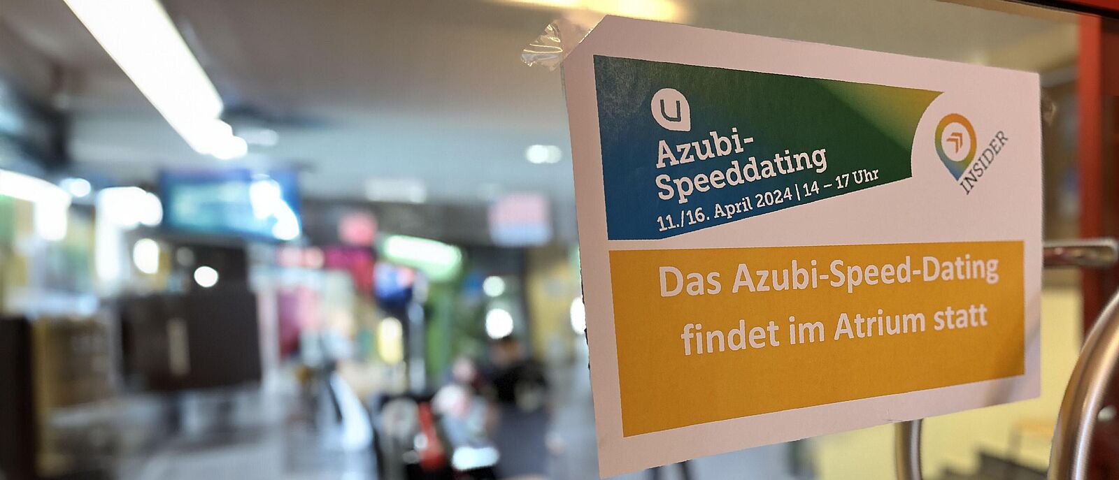 Auf einer Glastür mit Blick auf einem Warteraum ist ein A4 Blatt befestigt mit der Aufschrift \"Azubi-Speed-Dating findet im Atrium statt\"