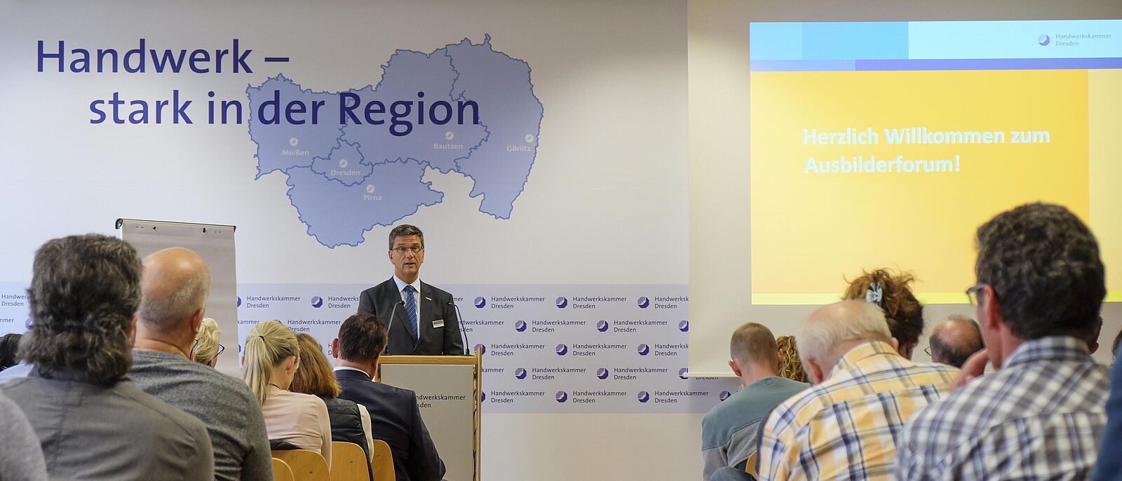 Blick in dem Konferenzraum über die Zuhörer Richtung Redner. Hinter ihm eine Präsentationswand.