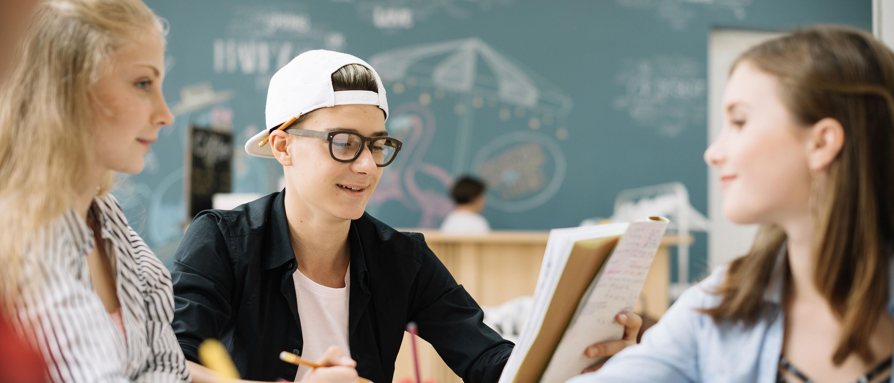 § Schüler sitzen gemeinsam am Tisch und lösen Aufgaben im Hintergrund eine Kreidetafel.