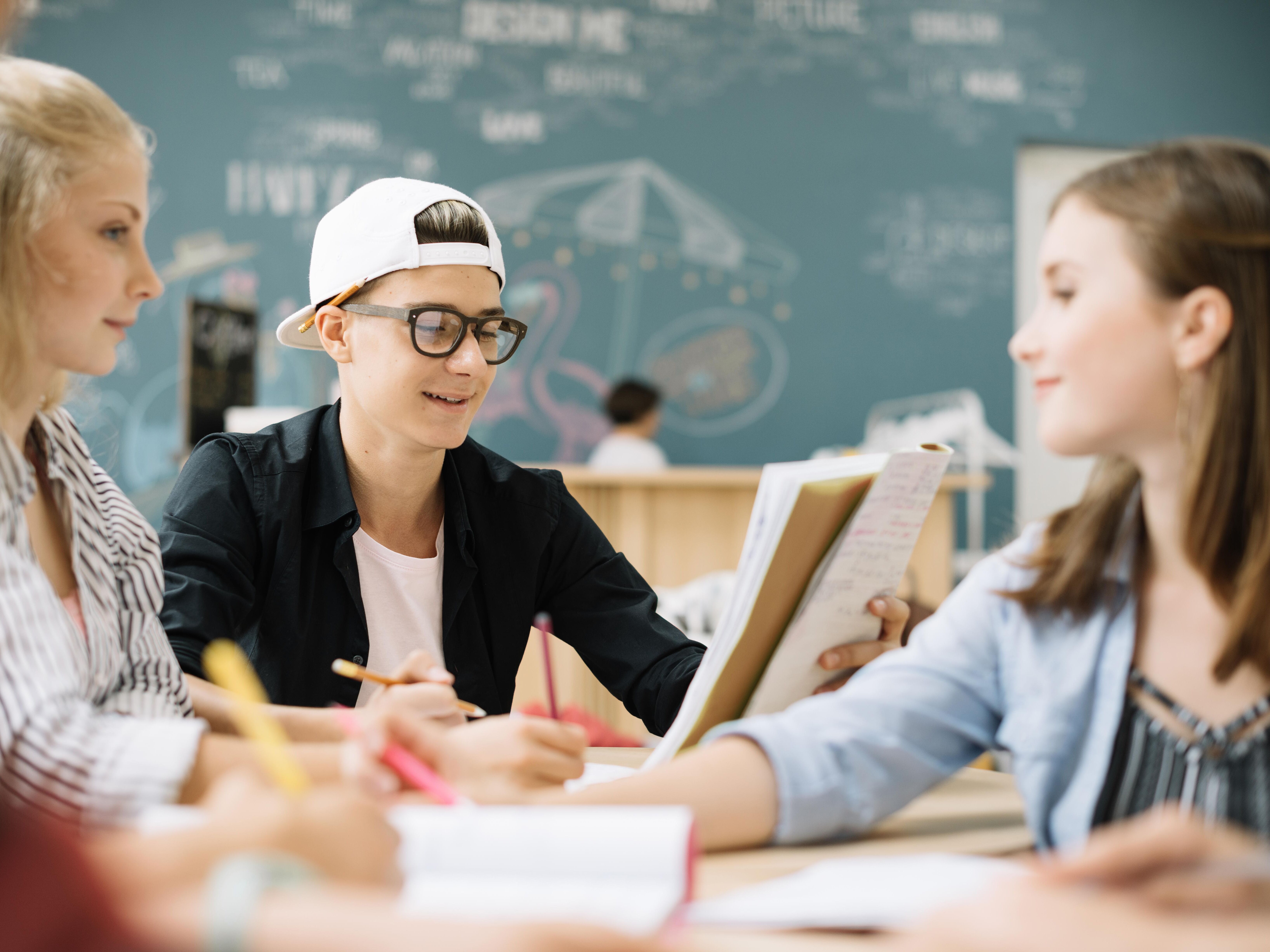 § Schüler sitzen gemeinsam am Tisch und lösen Aufgaben im Hintergrund eine Kreidetafel.