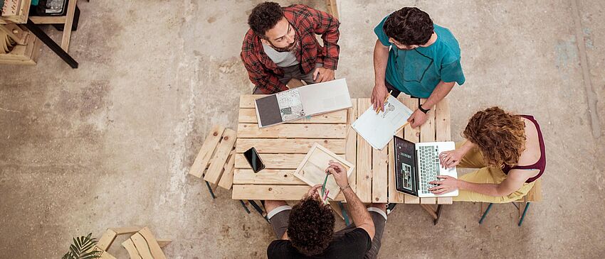 Macher-Mittwoch: Jeden letzten Mittwoch im Monat – Handwerkstreff im Telux Blick von oben: Vier Personen sitzen an einem Holztisch in einer Werkhalle und erarbeiten gemeinsam etwas. Eine Person recherchiert am Laptop. Die anderen diskutieren miteinander und machen sich auf den ausgelegten Dokumenten Notizen.