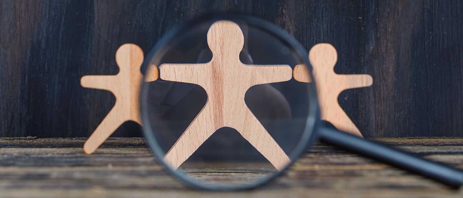 Auf einem Holztisch stehen drei Holzfiguren, darüber gelegt eine Lupe.