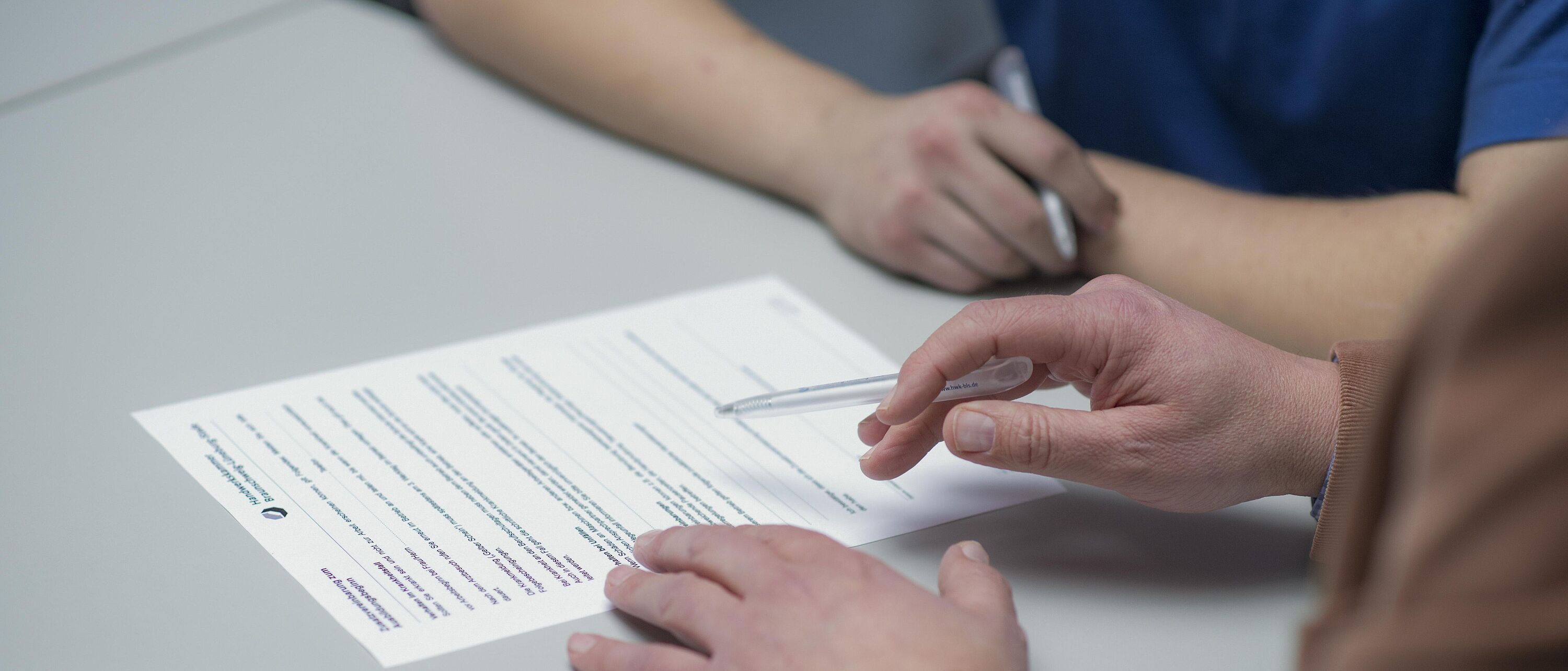 Makroaufnahme: Zwei Personen sitzen an einem Schreibtisch, vor sich einen Vertrag den eine Person unterschreiben will während die andere zeigt wo das Unterschriftenfeld ist.