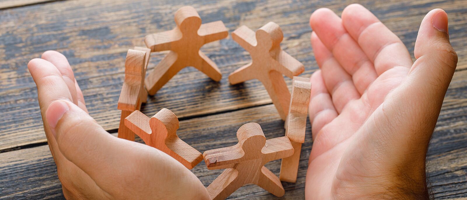 Auf einem Holztisch sind mehrere Holzfiguren ,die sich an den Händen halten und damit einen Kreis bilden. Darum zwei Hände die die Holzfiguren schützend umfassen.