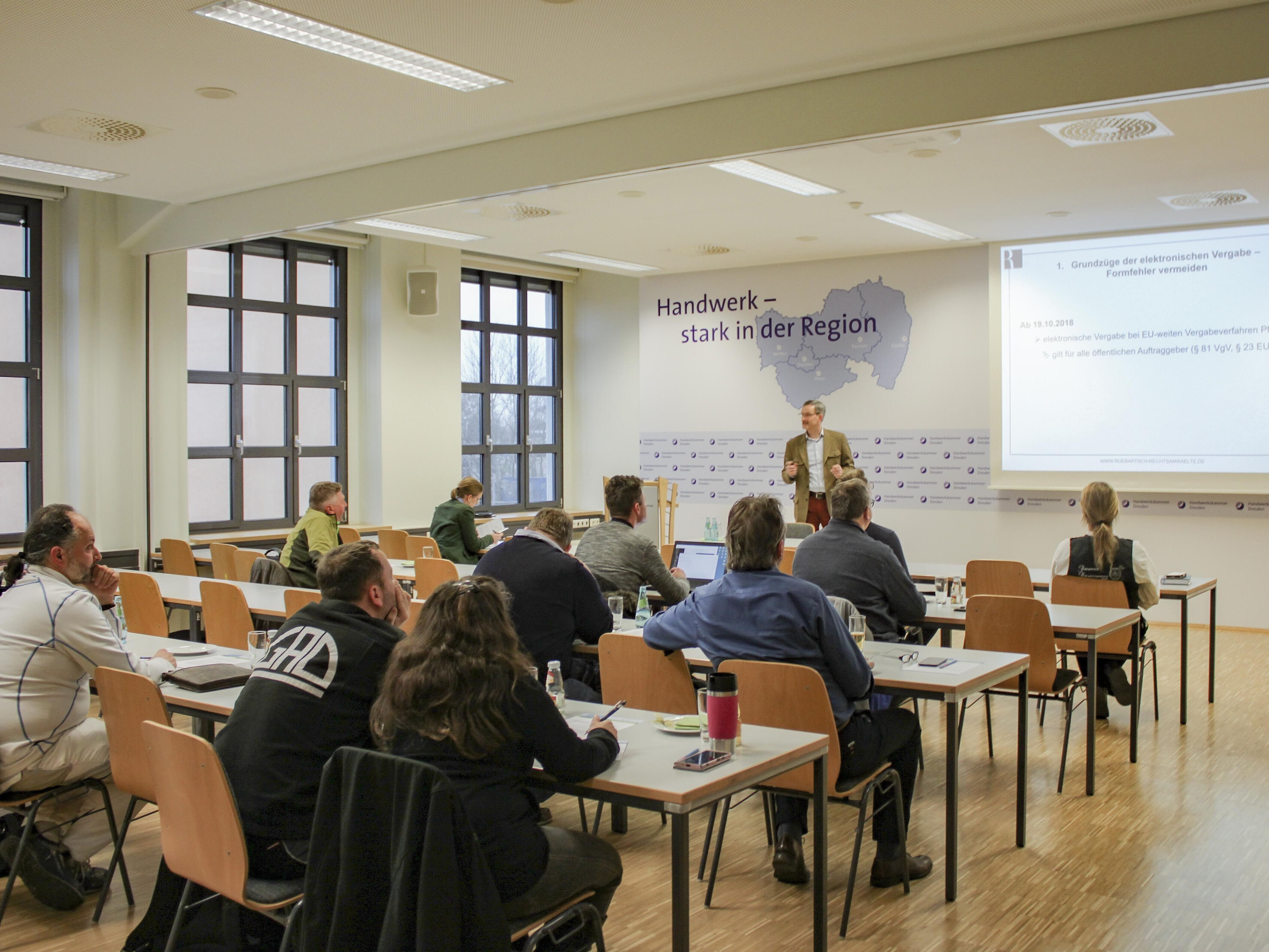 Blick in einem Veranstaltungsraum der Handwerkskammer Dresden Richtung Präsentations-Leinwand. Ein Referent informiert vor sich sitzende Zuhörer.