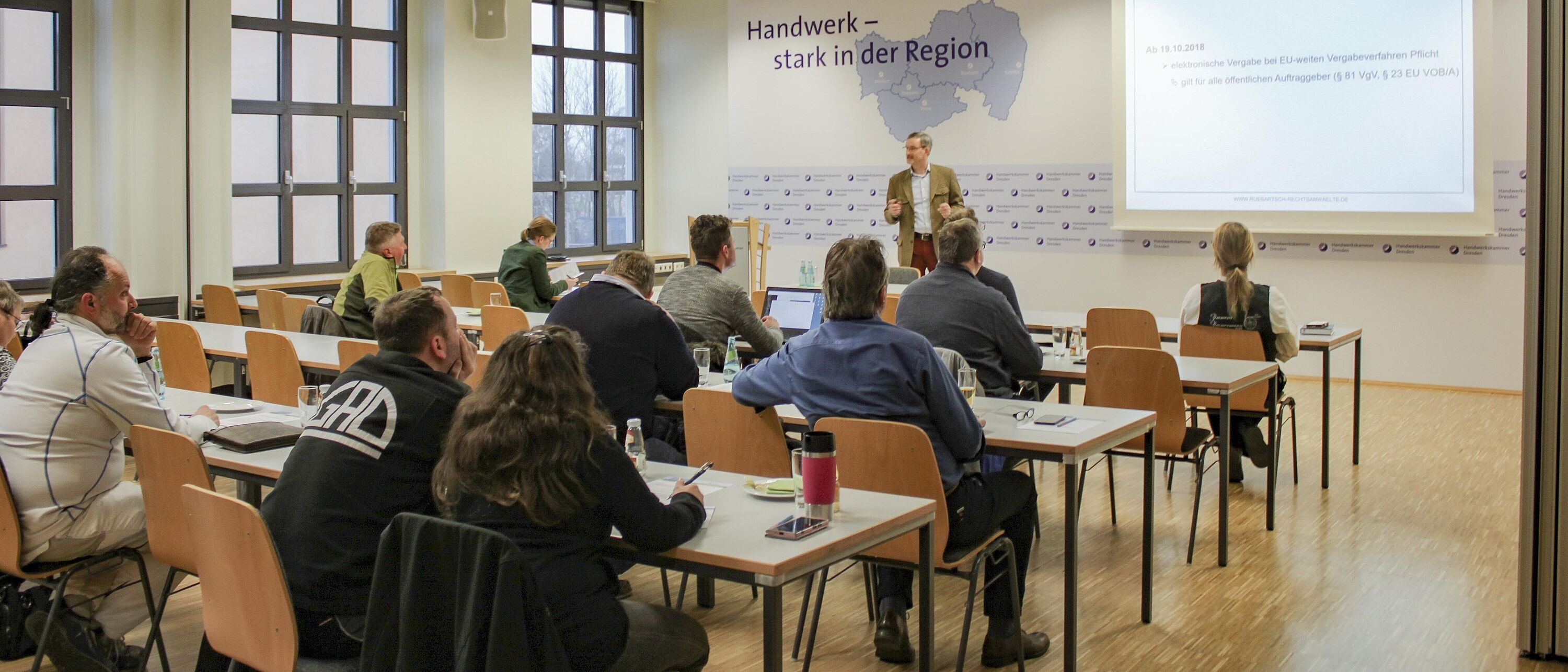Blick in einem Veranstaltungsraum der Handwerkskammer Dresden Richtung Präsentations-Leinwand. Ein Referent informiert vor sich sitzende Zuhörer.