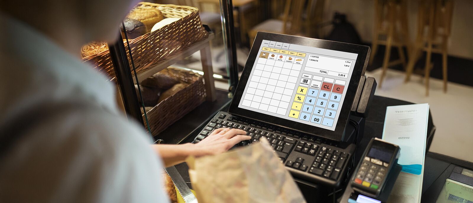 Eine Person steht hinter der Verkaufstheke vor einer elektronischen Kasse und hält in der rechten Hand eine Papiertüte. Mit der linken tippt sie gerade den Preis ein.