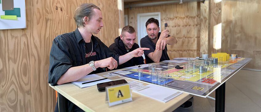 Drei Personen sitzen an einem Tisch und diskutieren kreativ. Vor ihnen liegt ein Planspiel auf einer Glasplatte. Darauf und daneben befinden sich abstrakte Spielfiguren, Klebezettel, Stifte sowie Anleitungs- und Dokumentationskarten.