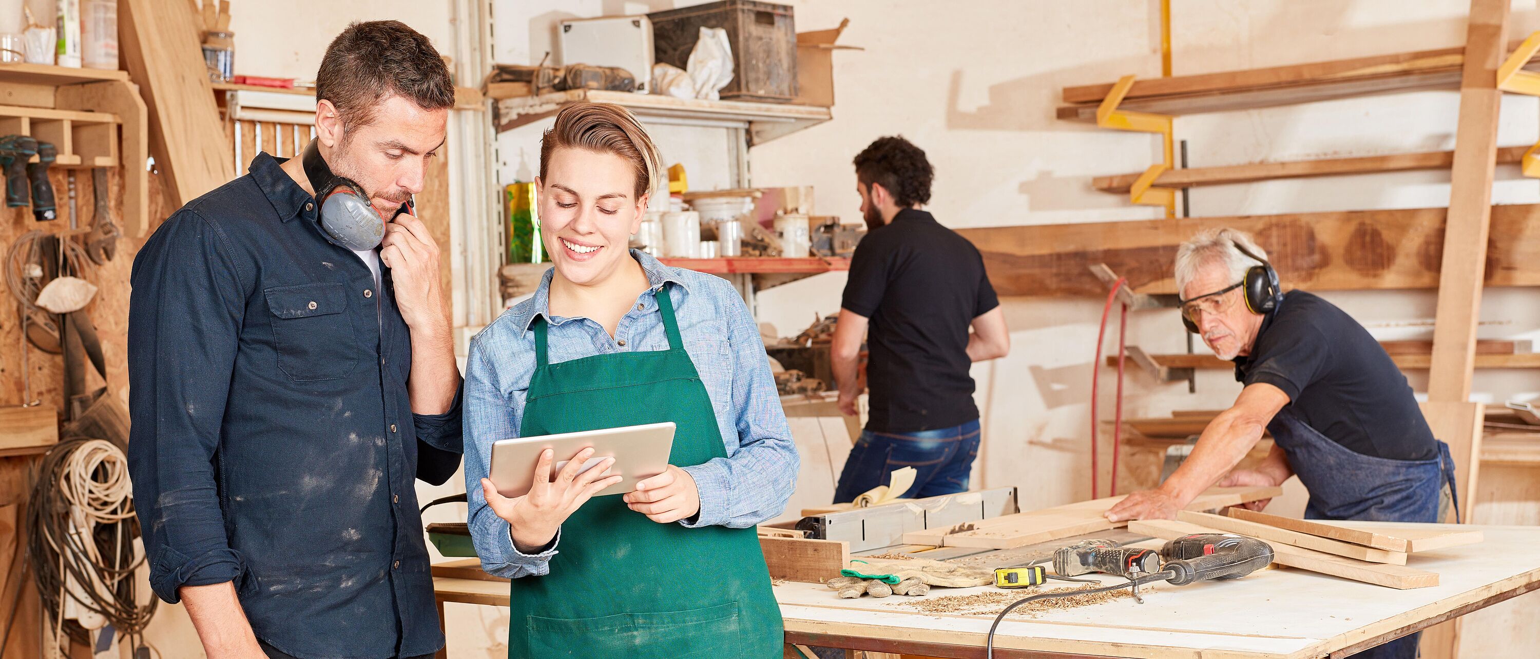 Zwei Personen stehen in einer Holzwerkstatt und schauen sich am Laptop, das eine Person hält, gemeinsam etwas an