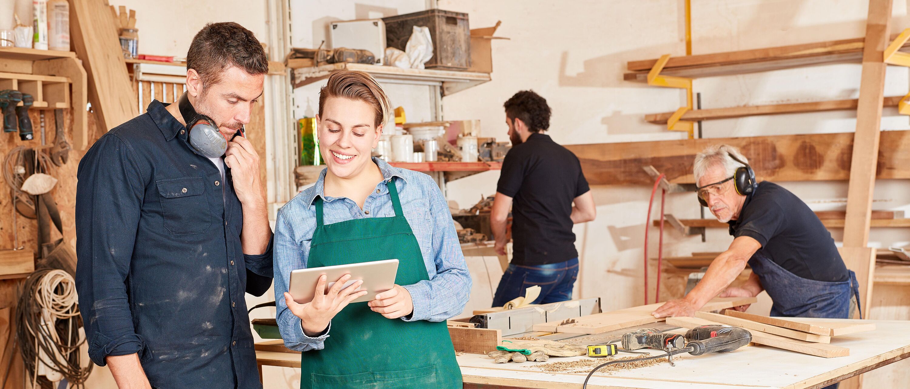 Zwei Personen stehen in einer Holzwerkstatt und schauen sich am Laptop, das eine Person hält, gemeinsam etwas an