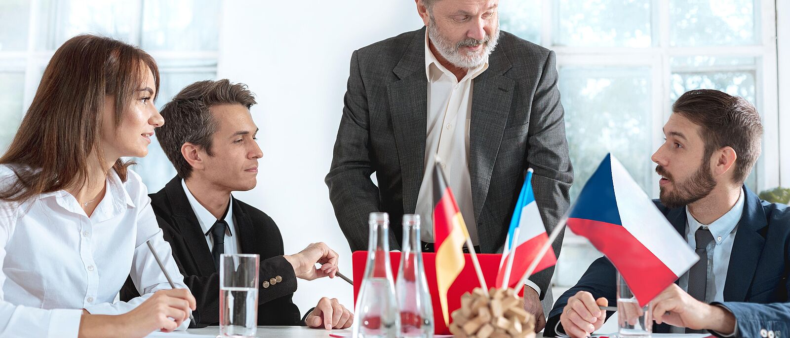 3 Personen sitzen und 1 Person steht am Tisch und beraten, in der Tischmitte stehen 3 Flaggen (Deutschland, Frankreich, Tschechien)