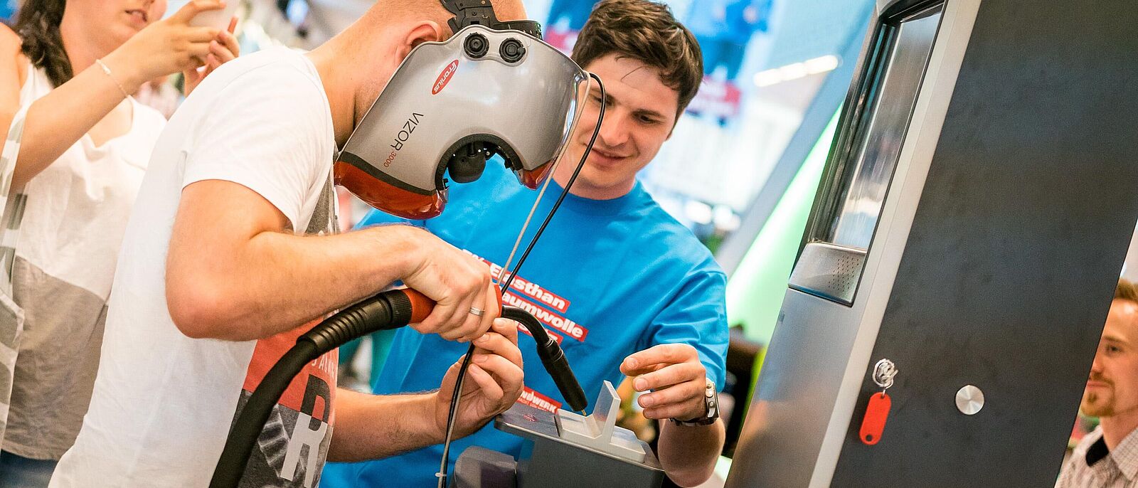 Eine Person hat eine virtuelle Schweißmaske auf und in der Hand ein virtuelles Schweigerät, damit steht er vor einem Schweißautomaten und versucht virtuell zu schweißen. Eine Person, die dahinter steht assistiert ihm dabei.