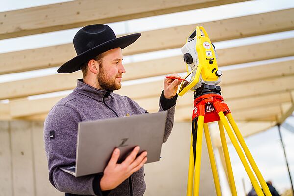 Eine Person mit einem Zimmerer-Hut benutzt ein modernes Tachymeter zum Ein- und Aufmessen auf der Baustelle.