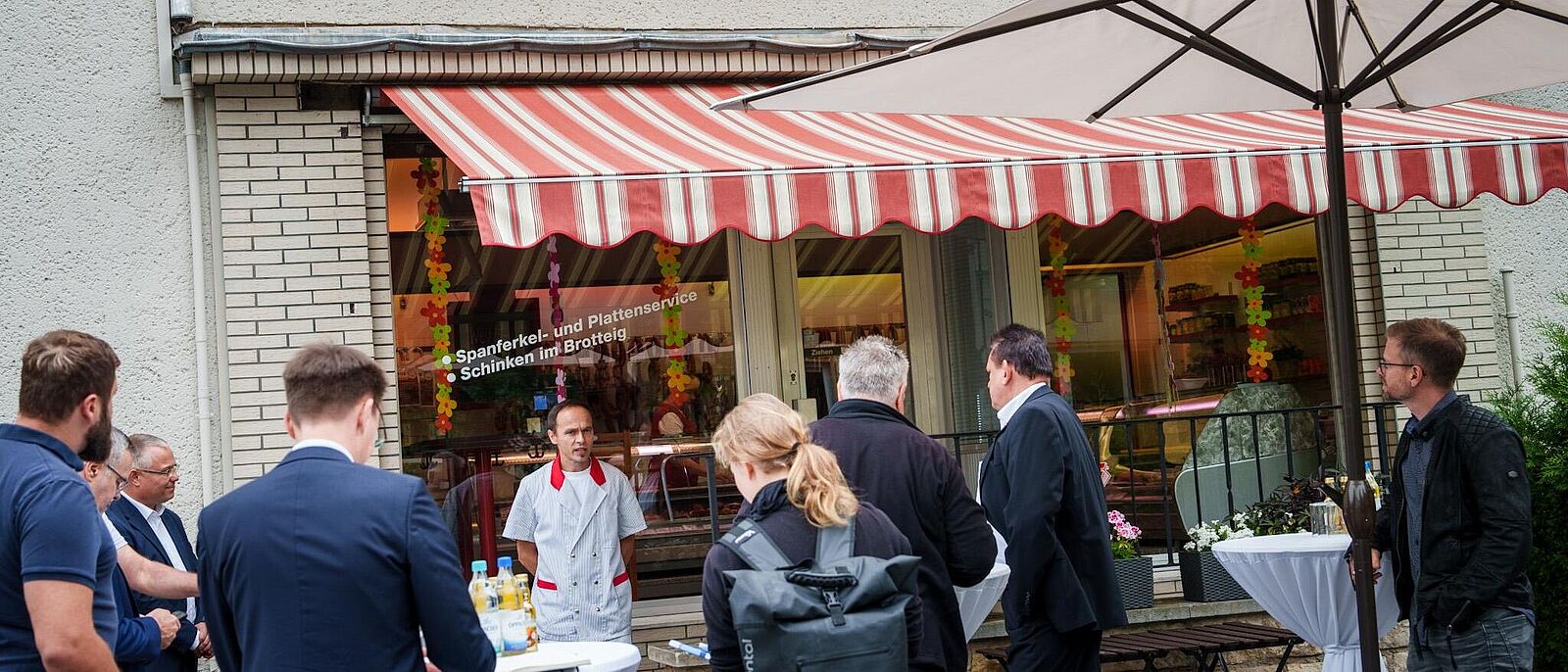 An der Station der  Pressetour: Fleischer W. Richter – Mehrere Personen der Tour stehen vor der Fleischerei bei einen kleinen Imbiss/Kostprobe während der Inhaber der Fleischerei über sein Geschäft informiert.