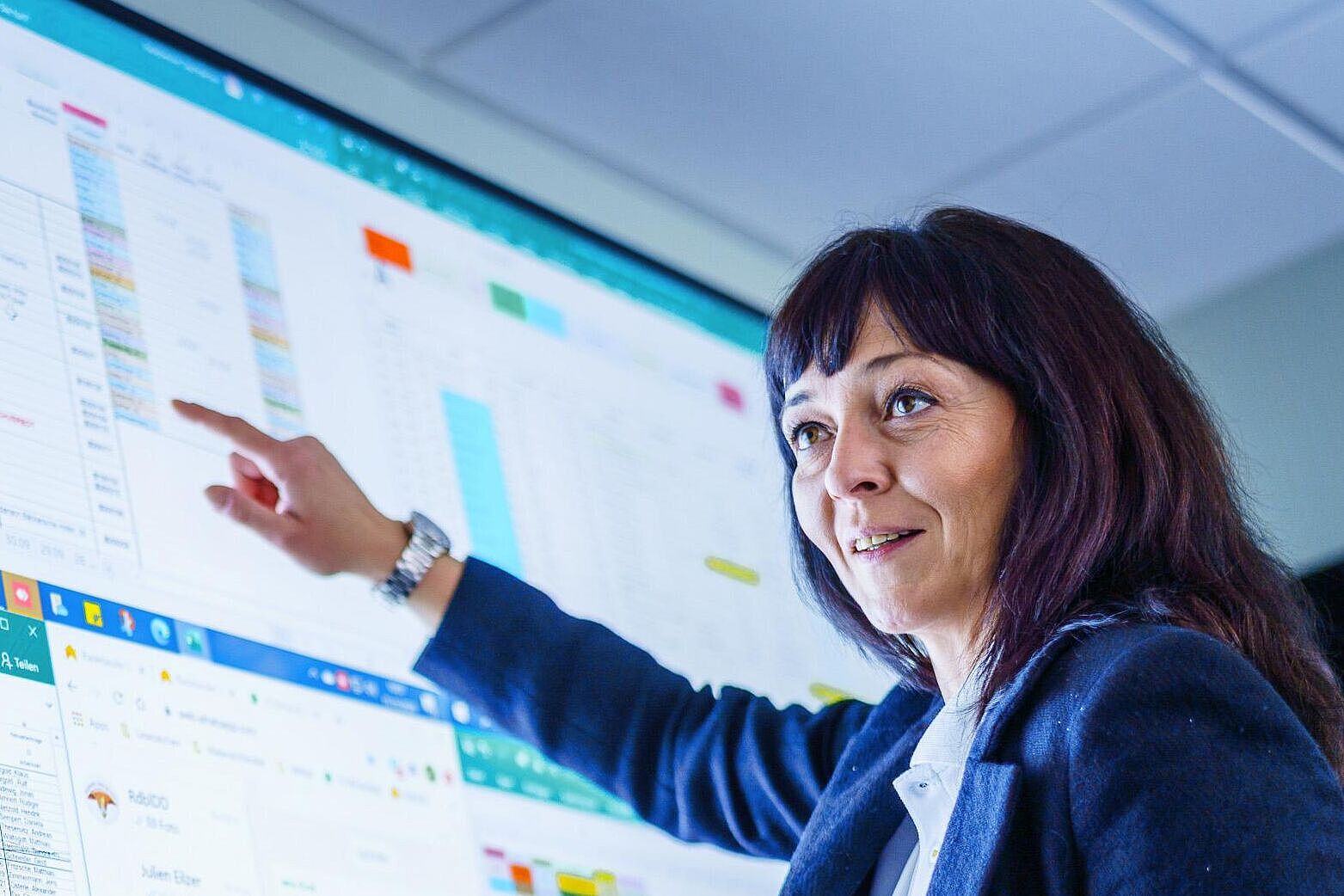 Digitaler Planungsbildschirm Zwei Personen stehen vor einem digitalen Planungsschirm. Die linke Person hält ein Handy in der rechten Hand. Die rechte Person zeigt mit der rechten Hand auf den Monitor und erklärt der anderen Person etwas.