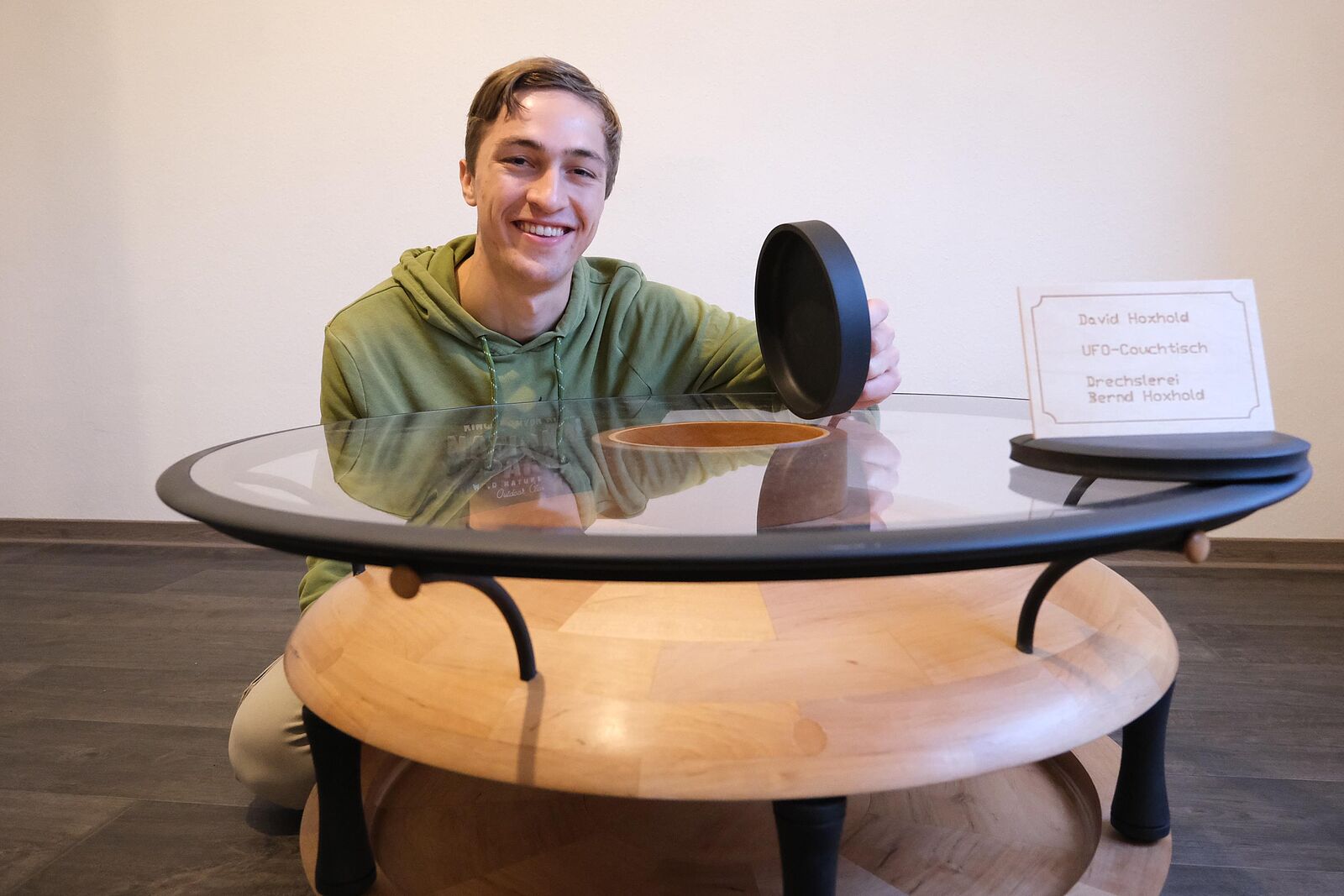 Ein Tischlergeselle hockt vor seinem Meisterstück \"Ufo-Couchtisch\", einem Couchtisch aus Holz mit hochgesetzer Glasplatte.