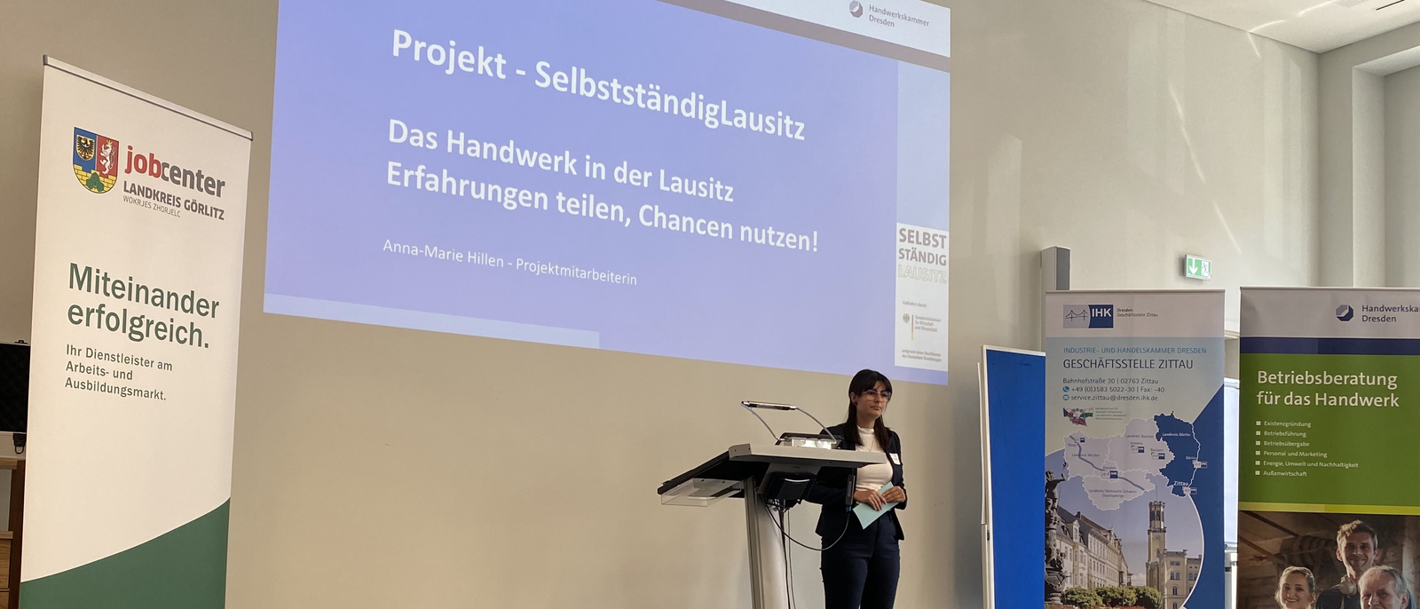 Blick auf eine Vortragende in einem Konferenzraum, daneben verschiedene Banner und Aufsteller, im Hintergrund eine Präsentation