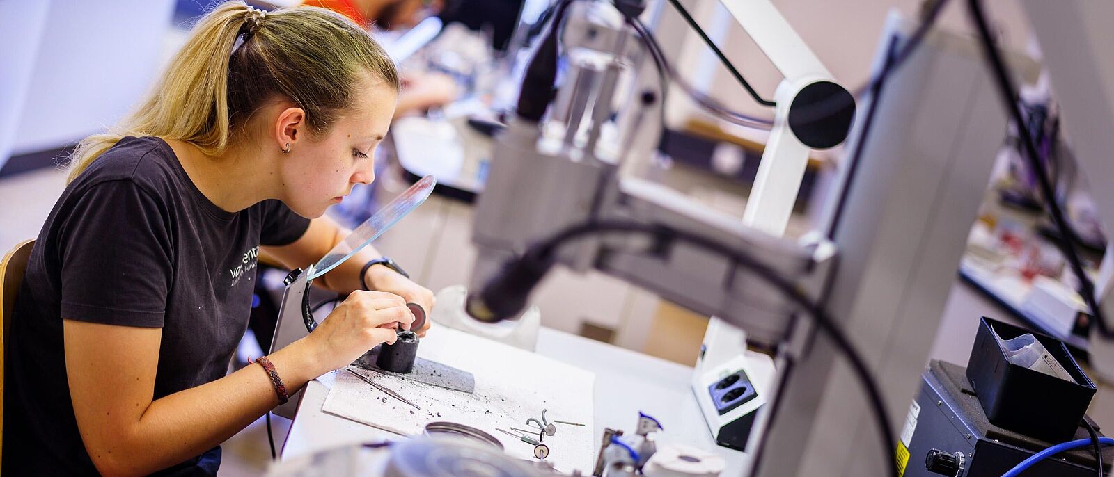 Eine Person sitzt an einem Arbeitsplatz mit Elektroanschluss und schaut dabei durch ein an der Werkbank angeschraubtes Vergrößerungsglas und reinigt oder schleift ein Werkstück.