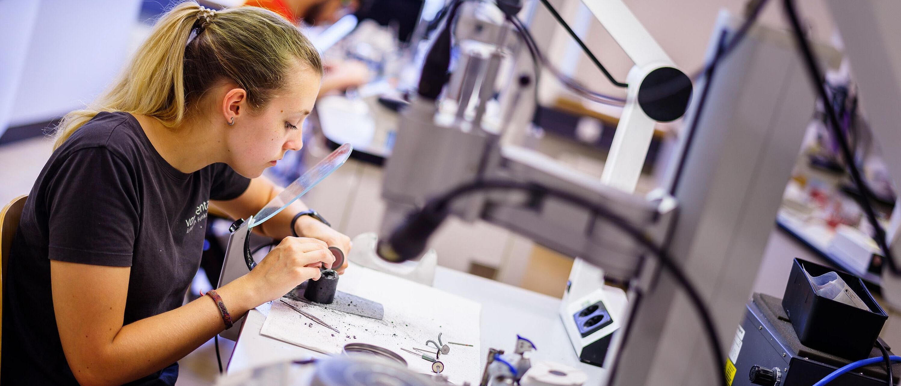 Eine Person sitzt an einem Arbeitsplatz mit Elektroanschluss und schaut dabei durch ein an der Werkbank angeschraubtes Vergrößerungsglas und reinigt oder schleift ein Werkstück.