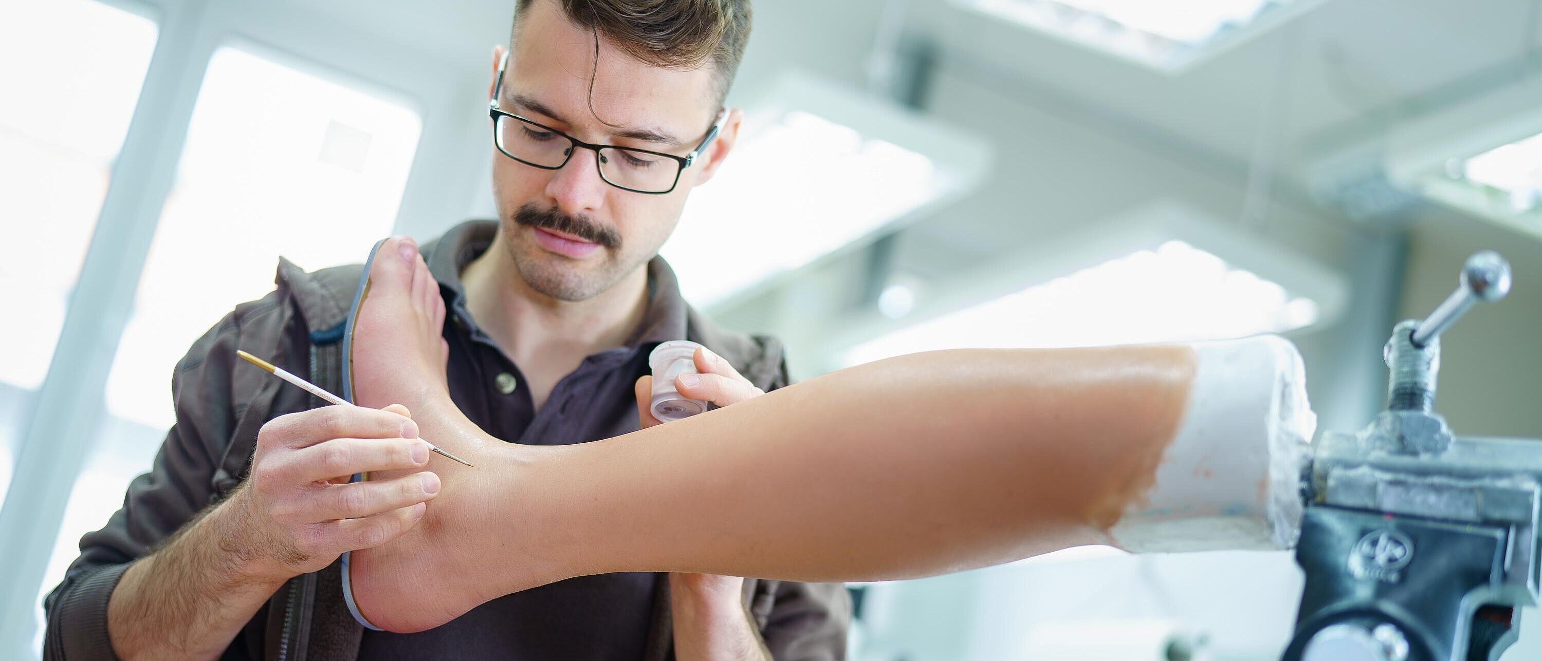 Eine Person, ein Orthopädietechniker bearbeitet mit einem Pinsel ein Silikonunterschenkel, welches in einen Schaubstock befestigt ist.