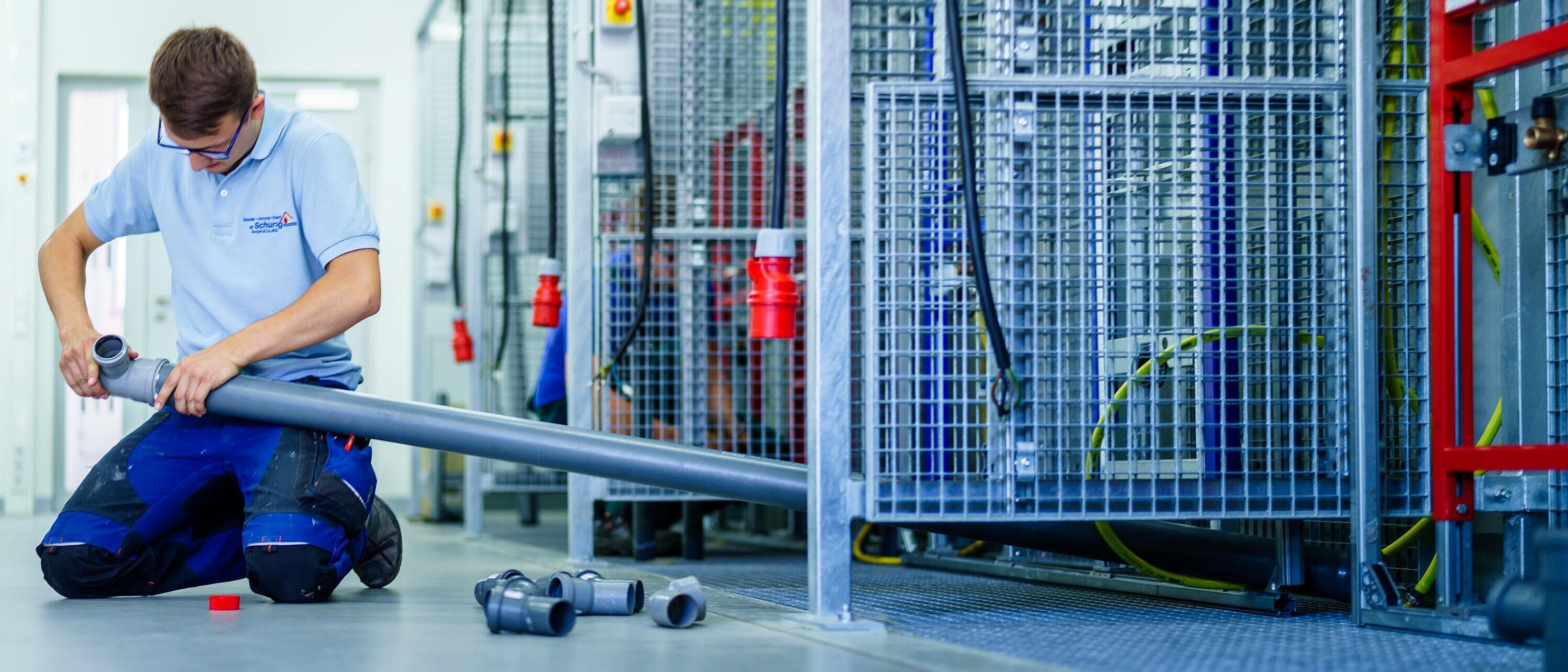 In einer Werkhalle vor einer praktischen Arbeitswand kniet eine Person und bearbeitet ein längeres Rohrstück.