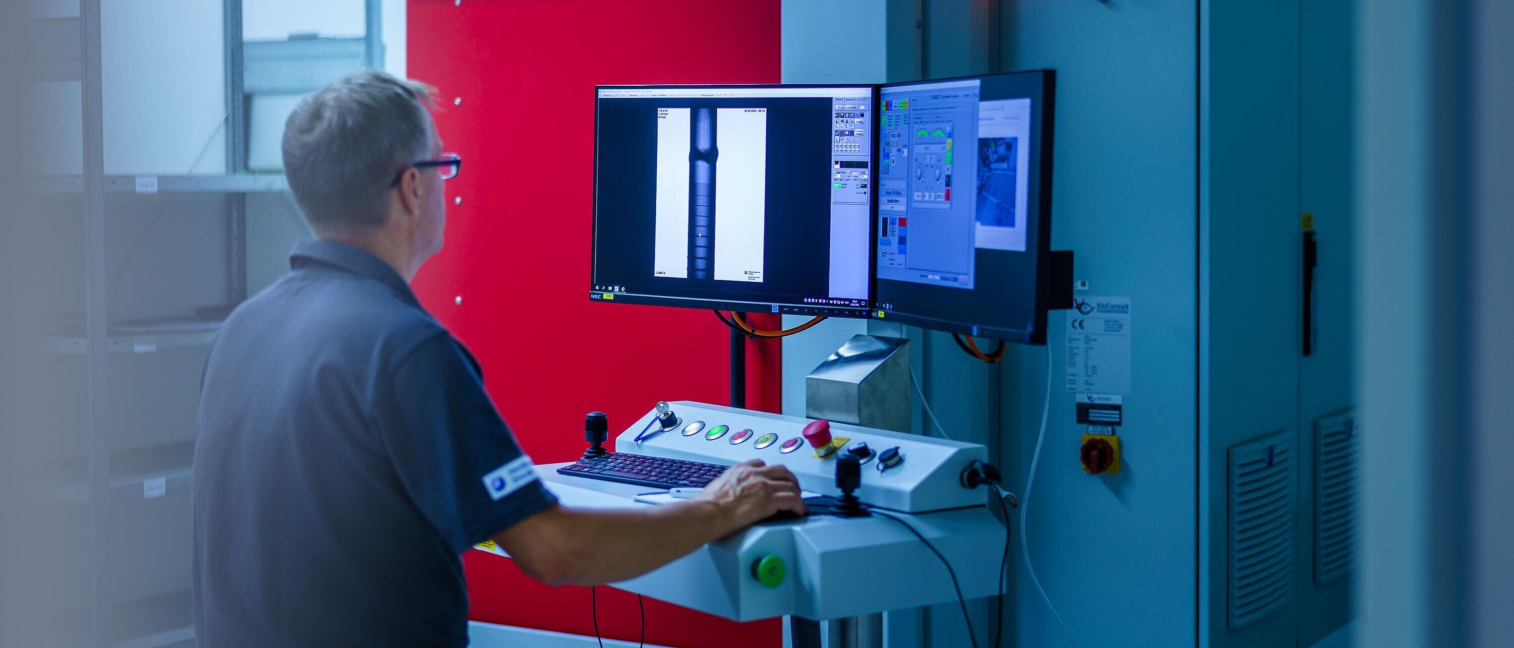 Eine Person steht vor einem großen Prüfschrank mit Bedienpult und zwei Monitoren, die Schweißnähte darstellen, und prüft diese.