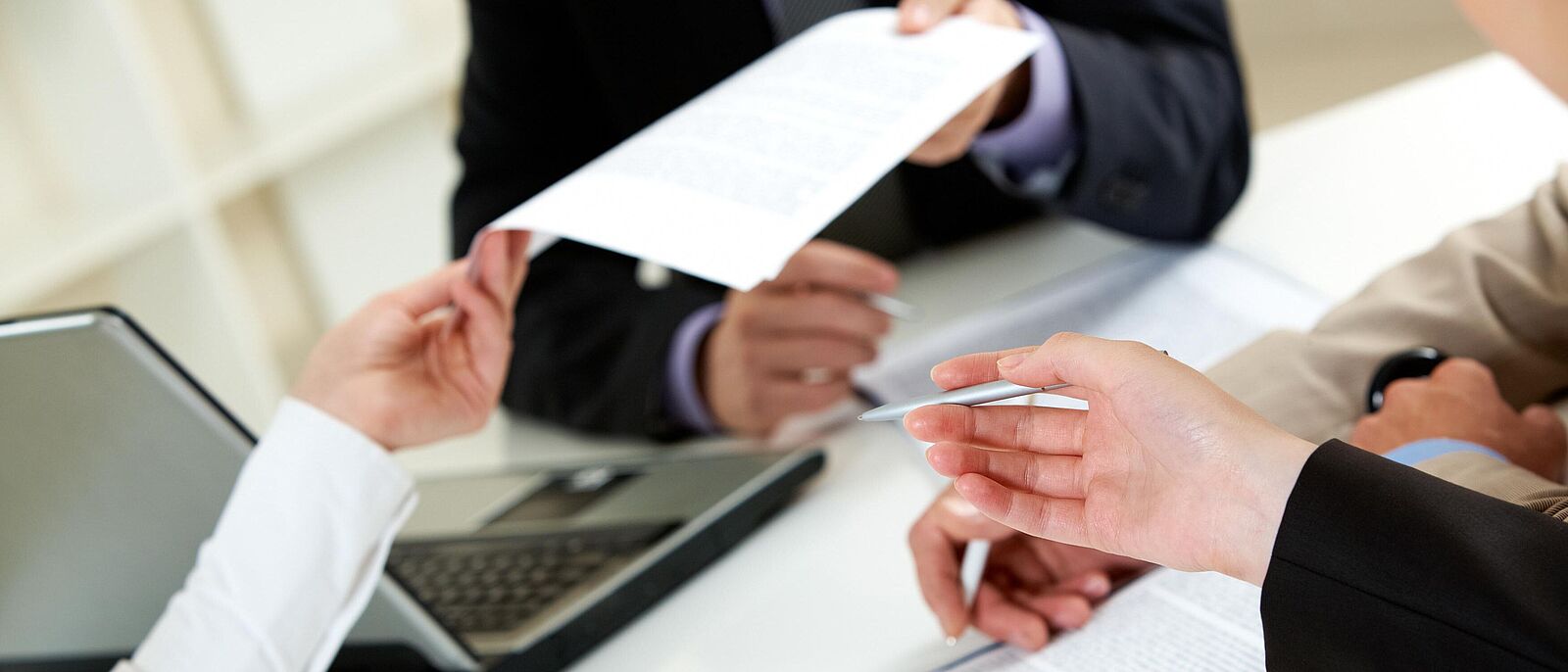 Drei Personen sitzen an einem Beratungstisch und tauschen sich Dokumente aus. Auf dem Schreibtisch befinden sich ein Laptop und verschiedene Dokumente.