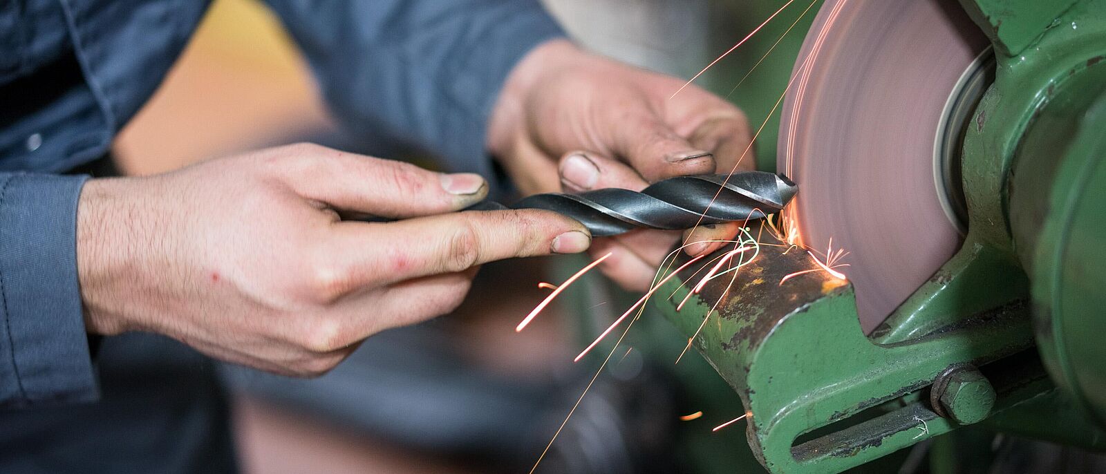 Eine Person in Arbeitskleidung eines Feinwerkmechanikers steht vor einer Werkbankmaschine und schleift ein Werkstück.