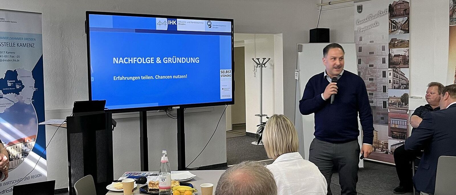 Eine Person steht mit einem Mikrofone vor einem Flipchart und informiert Interessierte, die beim Frühstück an kleinen Tischen sitzen.