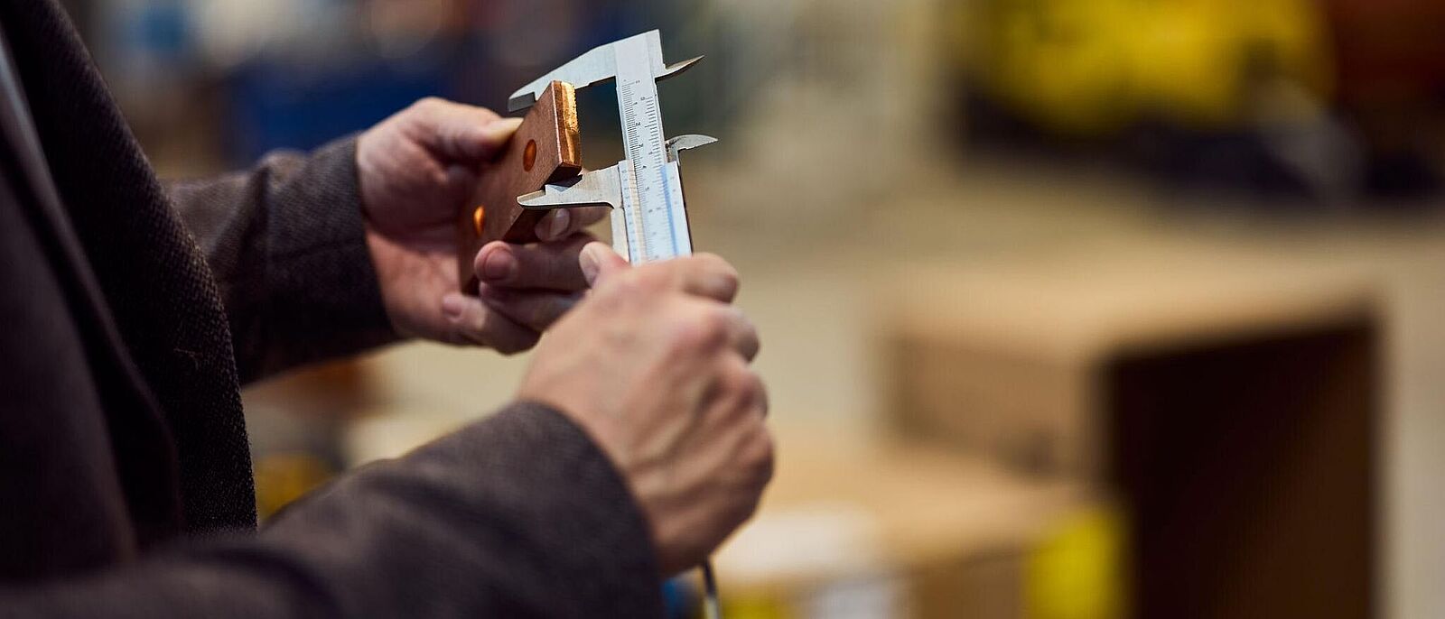 Handwerkergespräch: Normungen im Handwerk Es handelt sich um eine Macroaufnahme der Hände einer Person, die einen metallischen Werkstoff mit einem Messchieber prüft.