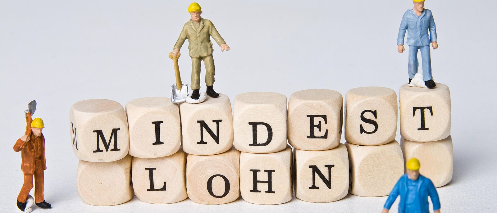 Grafik: Scrabbel- bzw. Holzsteine auf zwei Reihen übereinandergelegt, zu lesen ist das Wort  \"Mindestlohn\". Auf den Holzsteinen, daneben und davor Miniaturfiguren die verschieden Berufe darstellen.