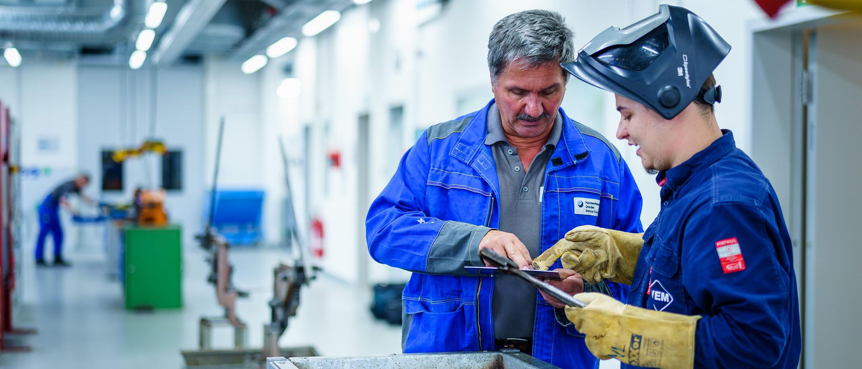 Ein Ausbilder und ein Lehrling stehen in einer Werkhalle vor einer Art Schweißmaterialcontainer und sehen sich ein Werkstück prüfend an.